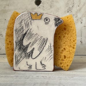 Può includere: Un portaspugna in ceramica bianca con un disegno di uccello, con una corona gialla e dettagli schizzati in nero. Il supporto è abbinato a una spugna gialla, creando un accessorio da cucina funzionale e decorativo.