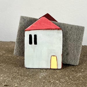 May include: A white ceramic house-shaped sponge holder with a bright red roof and a yellow door. The front facade features two vertical rectangular windows. A grey abrasive scouring pad is positioned behind the ceramic house structure.
