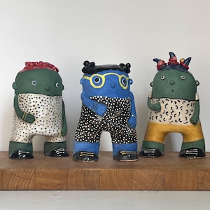 Könnte beinhalten: Drei Keramikfiguren stehen nebeneinander. Die linke Figur ist grün mit einem weiß gepunkteten Outfit und rotem Haar. Die mittlere Figur ist blau mit gelber Brille und schwarz gepunkteter Hose. Die rechte Figur ist grün mit einer gelben Hose.