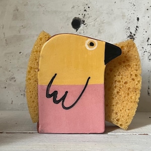 May include: A bird-shaped sponge holder with a yellow top and pink base, featuring a black beak and eye. Two yellow sponges are attached to the sides. This decorative item is designed to hold a kitchen sponge, adding a touch of fun to the sink area.