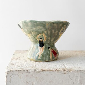 Puede incluir: Un cuenco de cerámica con borde acampanado y base de pedestal, con un esmalte verde salvia moteado. El exterior está decorado con figuras pintadas a mano de nadadores con bañadores negros y rojos, representados en varias posturas sobre el fondo.