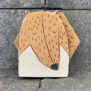 May include: A ceramic sponge holder shaped like a brown and white dog with closed eyes. The holder is sitting on a gray stone surface.