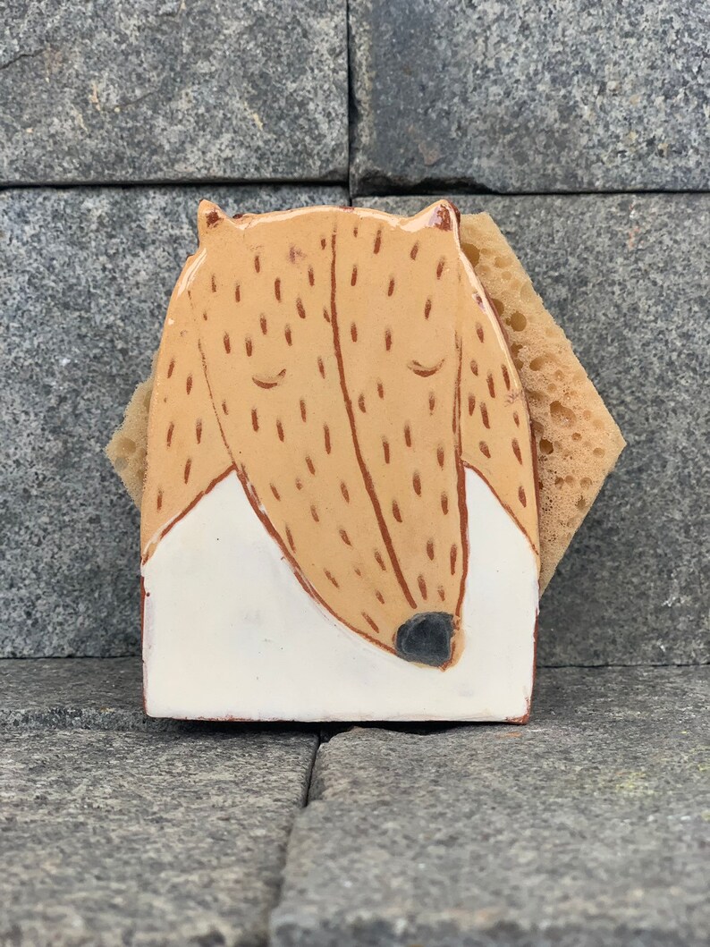 May include: A ceramic sponge holder shaped like a brown and white dog with closed eyes. The holder is sitting on a gray stone surface.
