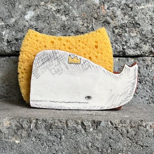 Peut inclure: Un porte-éponge en céramique blanc en forme de baleine avec une éponge jaune. La baleine a une couronne et un dessin esquissé. Le support a des accents rouges et repose sur une surface grise.