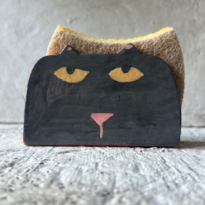 May include: A black ceramic cat-shaped sponge holder featuring yellow eyes, a pink nose, and small pointed ears. The holder contains a dual-textured yellow and brown cleaning sponge that extends slightly above the top edge of the cat figure.