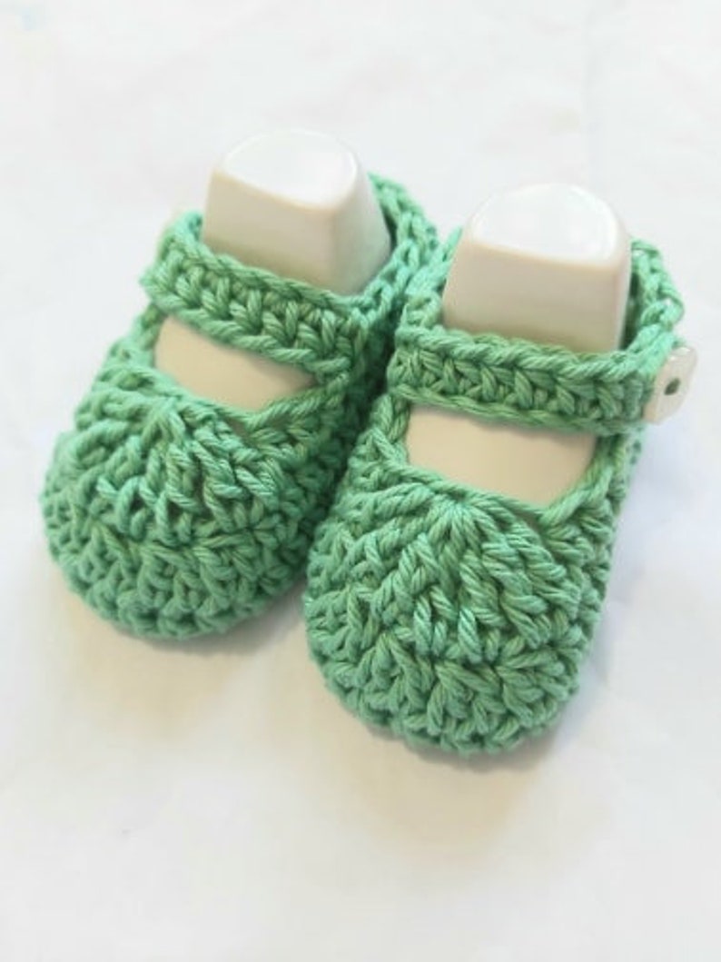 May include: A pair of green crocheted baby shoes with white buttons. The shoes are made with a simple stitch pattern and have a rounded toe.
