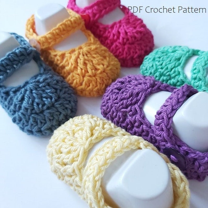 May include: Six pairs of crocheted baby shoes in different colors: blue, yellow, pink, green, purple, and cream. The shoes have a strap across the top and are displayed on white stands. PDF Crochet Pattern.