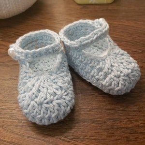 May include: A pair of light blue crocheted baby shoes with a pearl embellishment on each shoe.