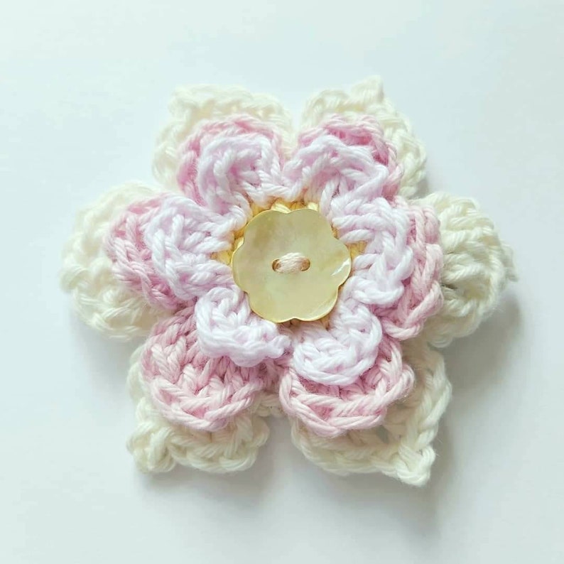 May include: A crocheted flower with pink and white yarn, featuring a pearl button in the center.
