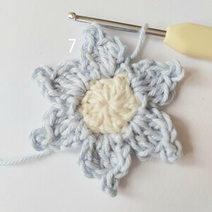May include: A light blue crochet flower with a white center. The flower is being worked on with a crochet hook and yarn. The number 7 is written on the image.
