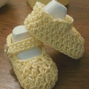 May include: A pair of crocheted baby shoes in a pale yellow color. The shoes have a simple design with a strap across the top.