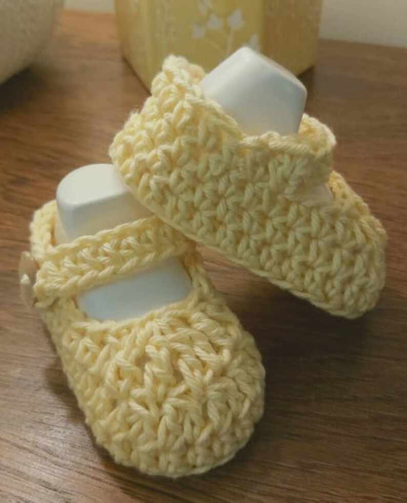 May include: A pair of crocheted baby shoes in a pale yellow color. The shoes have a simple design with a strap across the top.