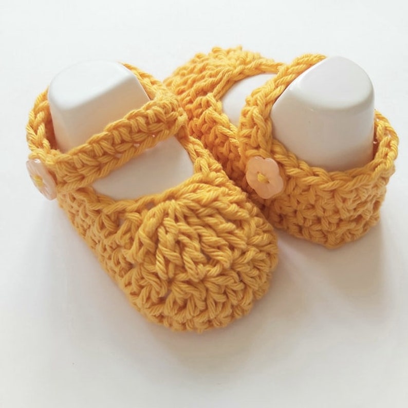 May include: A pair of yellow crocheted baby shoes with a flower button on each shoe.