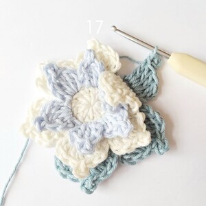 May include: A close-up of a crocheted flower with three layers in shades of blue and white. The flower is being worked on with a crochet hook and yarn.