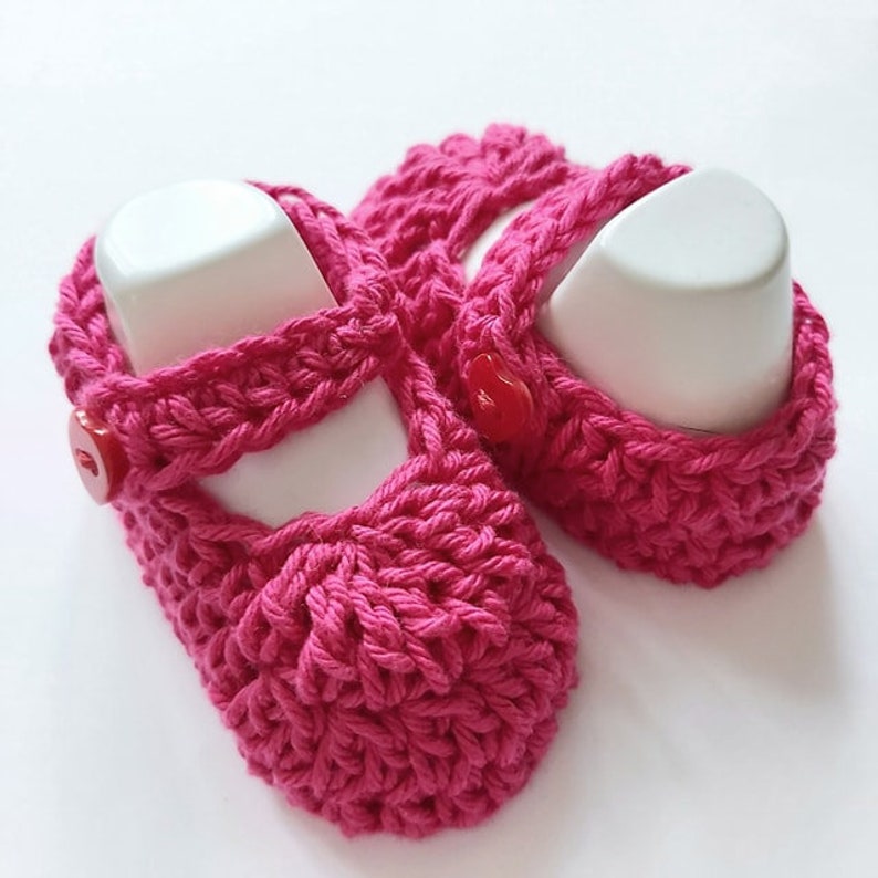 May include: A pair of pink crocheted baby shoes with red buttons. The shoes have a simple design and are perfect for a newborn.