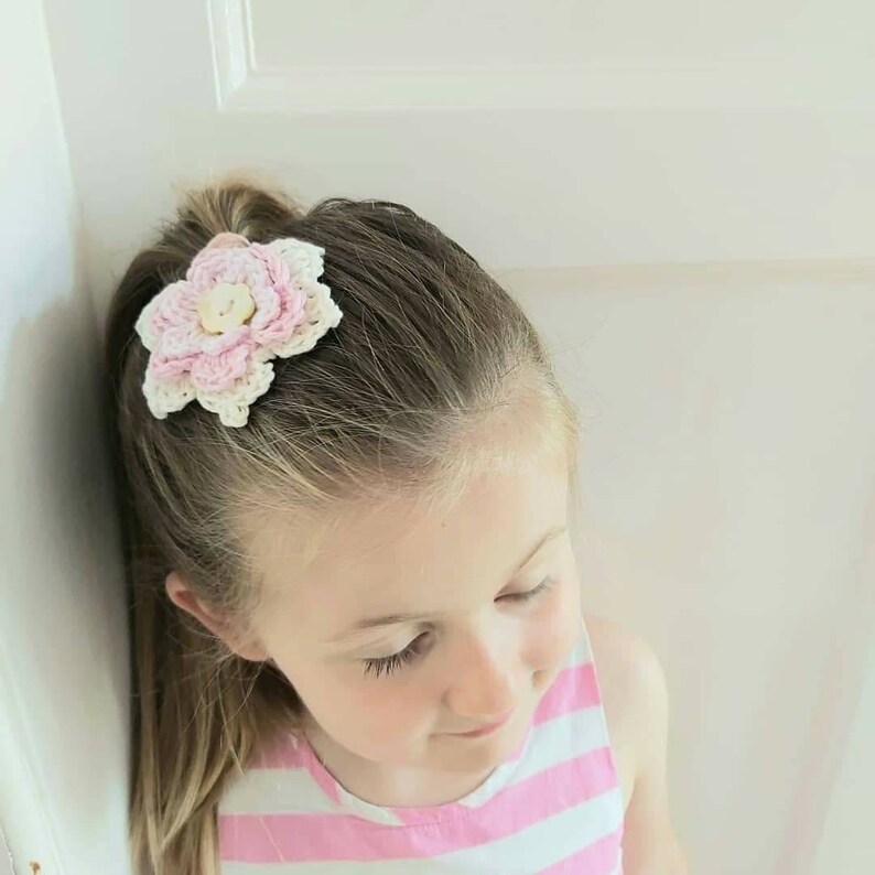 May include: A young girl with long brown hair wearing a pink and white striped shirt has a pink and white crocheted flower hair clip in her hair.