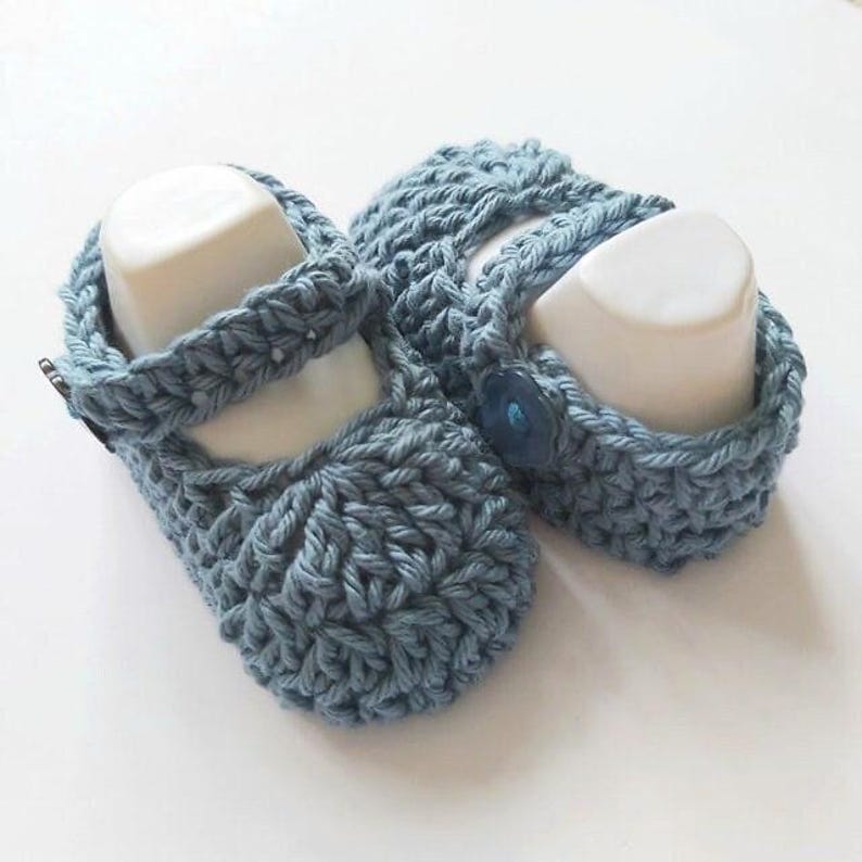 May include: A pair of blue crocheted baby shoes with a button detail. The shoes have a strap that goes around the ankle.