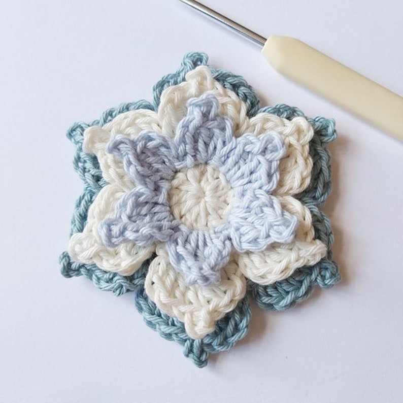 May include: A crocheted flower with three layers in shades of blue and white. The flower has six petals and is made with a delicate stitch.