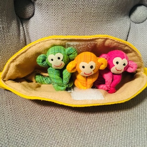 Può includere: Tre piccoli giocattoli di peluche a forma di scimmia in una borsa gialla a forma di banana. Le scimmie sono verde, arancione e rosa.
