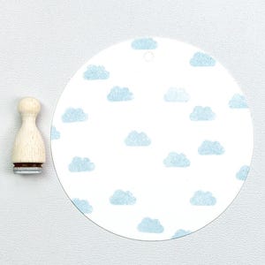 Op de afbeelding: Een houten stempel en een witte, ronde tag met een lichtblauw wolkenpatroon. De stempel heeft een houten handvat en een grijze basis. De tag heeft een klein gaatje aan de bovenkant.