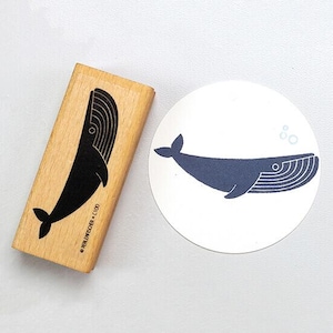 Könnte beinhalten: Ein Holzstempel mit einem schwarzen Wal-Design und dem Text "PERLENFISCHER C100" neben einem runden Aufkleber mit der gleichen marineblauen Wal-Illustration. Der Wal hat einen gebogenen Körper mit detaillierter Schwanzflosse und Linien auf dem Rücken.