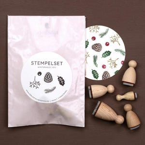 May include: A collection of wooden stamps with a winter theme, featuring pine cones and leaves, and a circular printed design. The stamps are packaged in a translucent bag with a label that reads "STEMPELSET WINTERWALD".
