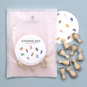 Op de afbeelding: Een set van negen kleine houten stempels met rubberen onderkant, verpakt in een doorschijnende roze papieren zak met een wit rond etiket met de tekst STEMPELSET ZAHLEN MINI S127 en kleurrijke cijfers van nul tot acht.