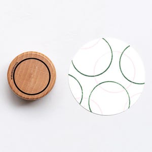 Op de afbeelding: Een houten flesstopper met een zwart ringontwerp en de tekst "HOLLENBECKER & CO." staat naast een witte onderzetter met een patroon van groene en roze overlappende cirkels. Op een witte achtergrond.