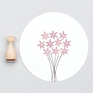 Könnte beinhalten: Ein Holzstempel mit Blumenmotiv neben einem weißen runden Anhänger mit einem rosa Blumenstrauß-Design. Der Stempel hat einen hellen Holzgriff und eine braune Basis. Der Blumenstrauß hat rosa Blütenblätter und schwarze Stiele.