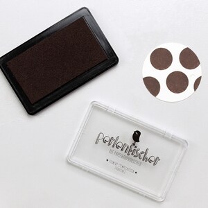 May include: A brown ink pad with a clear plastic case with the text "Perlenfischer Die Papierkrauts&uuml;chtigen" and a small bird illustration.