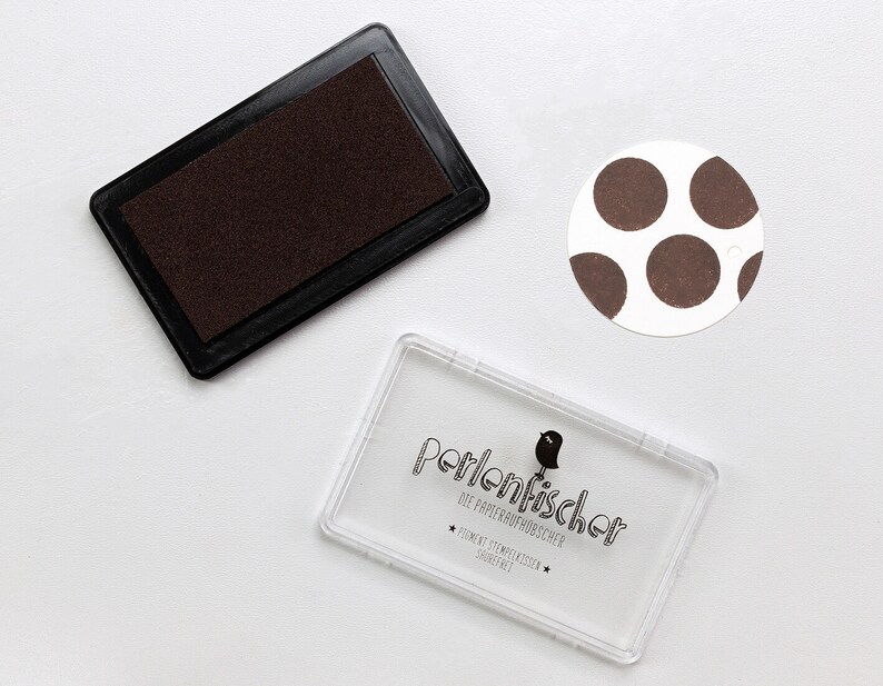 May include: A brown ink pad with a clear plastic case with the text "Perlenfischer Die Papierkrauts&uuml;chtigen" and a small bird illustration.