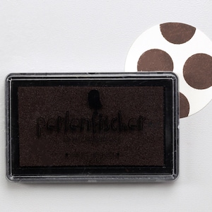May include: A brown ink pad with the text "Perlenschicker" on the front. The ink pad is in a clear plastic case. A white circle with brown polka dots is in the background.