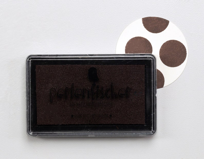 May include: A brown ink pad with the text "Perlenschicker" on the front. The ink pad is in a clear plastic case. A white circle with brown polka dots is in the background.