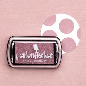 Può includere: Un tampone rettangolare con la scritta bianca "perlenfischer", un disegno di uccello e le parole "PIGMENT STEMPELKISSEN". Sullo sfondo un cerchio bianco con pois rosa. L'inchiostro è rosa.