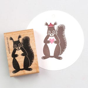 Puede incluir: Un sello de madera con un diseño de ardilla negra y el texto "PERLENFISCHER A253". Una imagen de ardilla a juego está impresa en una etiqueta blanca. La ardilla sostiene un corazón rosa y lleva un sombrero rojo.