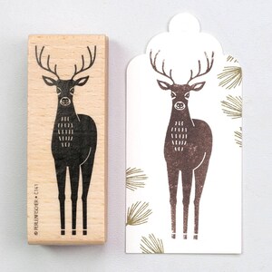 Könnte beinhalten: Ein Holzstempel mit einem schwarzen Hirschdesign neben einem bedruckten Etikett mit demselben Hirschbild. Der Stempel trägt den Text "PERLENFISCHER + C741". Das Etikett hat einen weißen Hintergrund mit grünen Tannenzweigen.