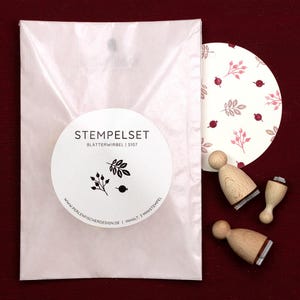 May include: A stamp set with a white package and a round card featuring a berry and leaf pattern. The set includes three wooden stamps with rubber designs. The package has a sticker with the text "STEMPELSET" and botanical illustrations.