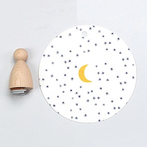 May include: A wooden stamp with a light brown handle and a circular base. Next to it is a white, circular tag featuring a yellow crescent moon and blue star illustrations. The tag has a small hole at the top.