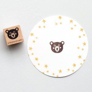Könnte beinhalten: Ein Holzstempel mit einem Bärengesicht-Design neben einem weißen runden Anhänger mit passendem Bärengesicht und goldenen Sternen. Der Stempel trägt den Text "PERLENFISCHER.BOIS" an der Seite.