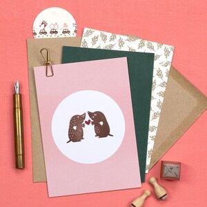 May include: A collection of stationery items on a pink background. Includes a pink card with a white circle featuring two brown moles holding a red heart, a gold pen, and a small stamp.