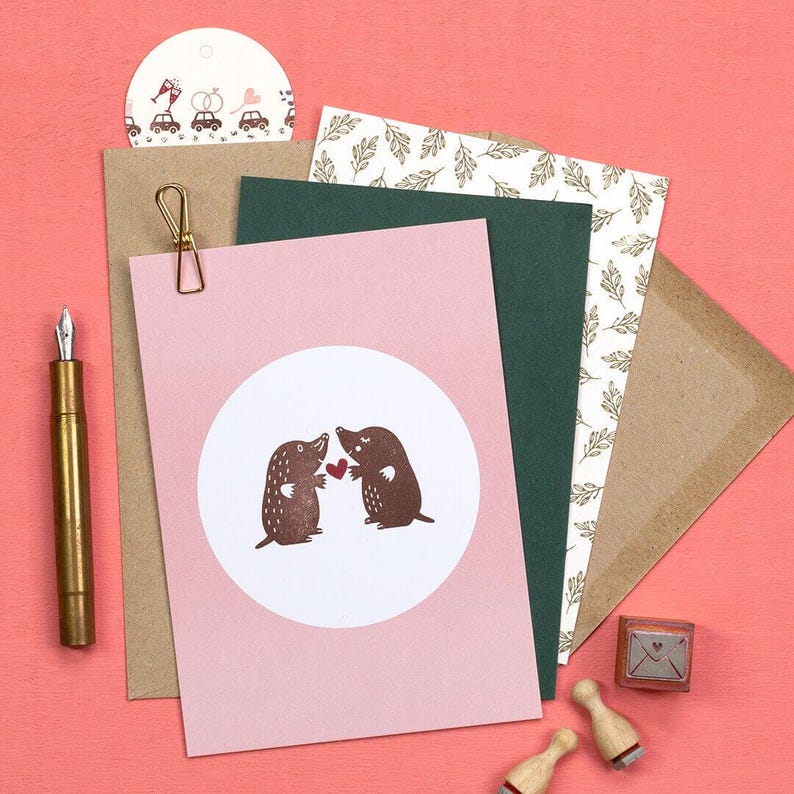 May include: A collection of stationery items on a pink background. Includes a pink card with a white circle featuring two brown moles holding a red heart, a gold pen, and a small stamp.