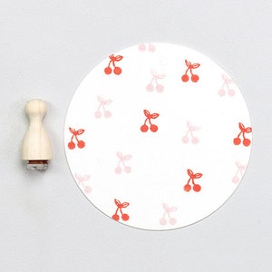 Puede incluir: Un sello de madera redondo con un diseño de cerezas. El sello es rojo y rosa y tiene un fondo blanco. El sello tiene un diámetro de aproximadamente 5 cm.