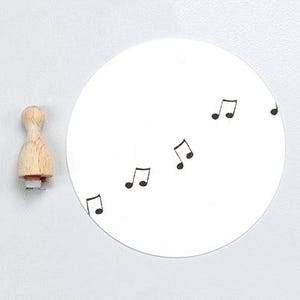 May include: A wooden stamp with a musical note design. The stamp has a light brown handle and a red base. A white circular paper has several black musical notes printed on it.