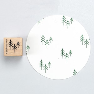 Puede incluir: Un sello de madera con un diseño de árbol de bosque junto a un papel circular blanco con un patrón repetido de árboles de bosque. El sello tiene el texto "PERLENFISCHER + B075".