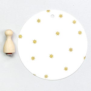 Pode incluir: Um carimbo de madeira com um design em forma de estrela e uma etiqueta circular branca com padrões de estrelas douradas com glitter. O carimbo tem um cabo de madeira clara e uma base vermelha.