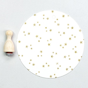 May include: A wooden stamp with a star design next to a white, circular tag patterned with gold stars. The stamp has a wooden handle and a red rubber base. The tag has a small hole at the top.