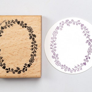 May include: A wooden stamp with a black floral wreath design. The stamp is rectangular and has a light brown colour. The floral wreath design is a circle with leaves and flowers.