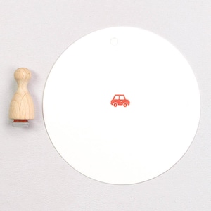 May include: A wooden stamp with a car design and a white circular tag with a red car graphic. The stamp has a natural wood handle and a red car image on the stamp base. The tag has a small hole at the top.