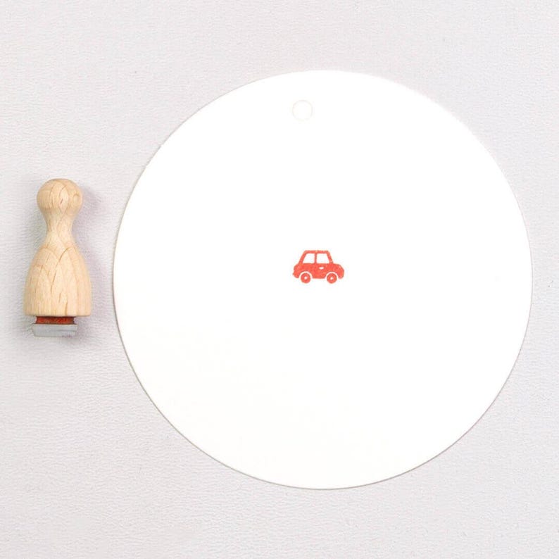 May include: A wooden stamp with a car design and a white circular tag with a red car graphic. The stamp has a natural wood handle and a red car image on the stamp base. The tag has a small hole at the top.