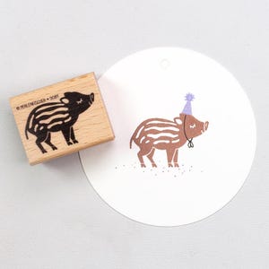 Könnte beinhalten: Ein rechteckiger Holzstempel mit einem schwarzen Stempelabdruck eines gestreiften Ferkels und dem Text PERLENFISCHER D084. Daneben liegt ein weißer, runder Papieranhänger mit einem braunen Abdruck eines Ferkels, das einen hellvioletten Partyhut trägt.